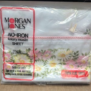 Puede incluir: Una sábana plana doble empaquetada de Morgan Jones. La sábana blanca presenta un estampado floral con margaritas rosas y amarillas. El embalaje incluye el texto "No-Iron luxury muslin sheet" y está diseñada para un colchón de 99 cm x 193 cm.