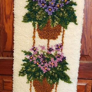 May include: A rectangular latch hook wall hanging featuring three stacked hanging baskets filled with purple and pink flowers and green foliage against a cream-coloured background. The piece is suspended from a wooden hanger with metal clips.