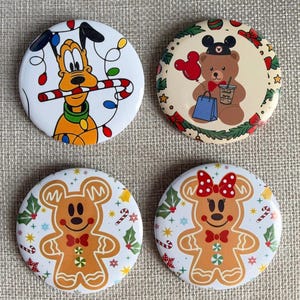 May include: Four circular pinback buttons featuring holiday designs. One shows Pluto with a candy cane and string lights, another a bear with a Mickey hat and shopping bag, and two show gingerbread bears with holly and candy canes.