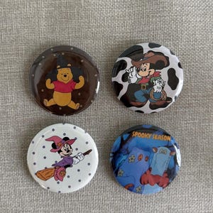 May include: Four circular pinback buttons featuring Disney characters. One shows Winnie the Pooh in a witch hat, one shows Mickey Mouse as a cowboy, one shows Minnie Mouse on a broomstick, and one shows Tigger and Eeyore with the text SPOOKY SEASON.