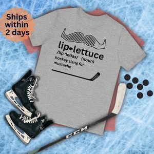 Puede incluir: Una camiseta gris con un palo de hockey negro y un gráfico de bigote. El texto en la camiseta dice "lip-lettuce /lip 'ledas/ (sustantivo) jerga de hockey para bigote".
