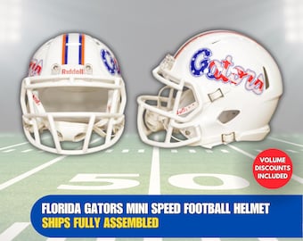 Op de afbeelding: Witte mini speed-voetbalhelm met een blauw en rood "Gators"-logo met sterren. De helm staat op een groen-wit voetbalveld.