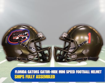 Op de afbeelding: Twee mini-American footballhelmen, één met het Florida Gators-logo en de andere met nummer 1. De helmen zijn donker metallic groen met zwarte gezichtsmaskers. De tekst op de afbeelding luidt: FLORIDA GATORS GATOR-HIDE MINI SPEED FOOTBALL HELMET.