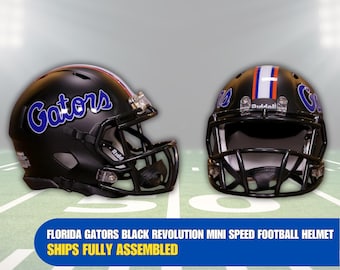 Op de afbeelding: Zwarte mini-voetbalhelm met een blauw en wit Gators-logo en een zwart gezichtsmasker. De helm staat op een groen en wit veld.