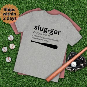 May include: A heather grey t-shirt with a baseball bat graphic and the definition of "slugger" printed on it. The text reads: "slug.ger /'sləgər/ (noun) a baseball player who commonly hits with great power."