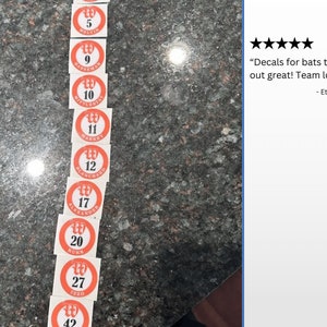 Pode incluir: Um conjunto de 10 adesivos de n&uacute;meros para taco de beisebol vermelho e branco com letras brancas. Os n&uacute;meros s&atilde;o 5, 9, 10, 11, 12, 17, 20, 27 e 42. Os adesivos est&atilde;o em uma bancada de granito.