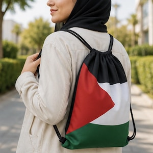 Op de afbeelding: Een rugzak met trekkoordsluiting voorzien van de horizontale zwarte, witte en groene strepen van de Palestijnse vlag met een rode driehoek aan de linkerzijde. De tas heeft zwarte koordhengsels en wordt gedragen over een lichtgekleurd jasje.