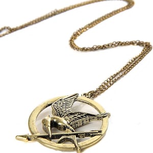 May include: A gold-toned necklace with a pendant featuring a stylized mockingjay bird in flight, with an arrow through its wings, inside a circular frame.