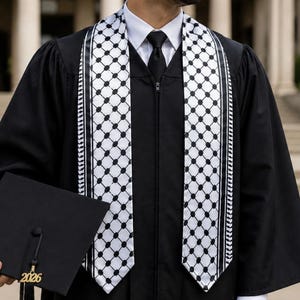 May include: A black graduation gown with a white shirt and black tie, worn with a black and white keffiyeh-patterned graduation stole. A black graduation cap with a gold 2026 tassel is held in the hand, completing the academic attire.