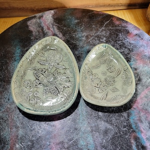 May include: Two ceramic dishes with a light green glaze and a floral design featuring a turtle. The dishes are on a black surface with blue and pink swirls.