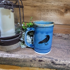 May include: A blue ceramic mug with a black raven illustration. The mug has a handle and a white base. The background includes a rustic wooden surface and a candle lantern.