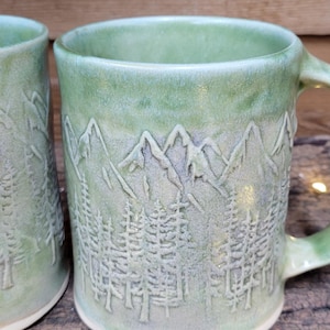 May include: Two ceramic mugs with a light green glaze featuring an embossed design of a mountain range above a dense forest of pine trees. The mugs have a smooth, glossy finish and a cylindrical shape with a sturdy handle on the side.