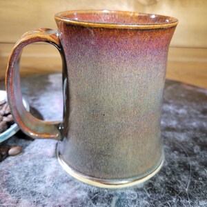 May include: A large, brown ceramic mug with a speckled glaze. The mug has a wide, rounded handle and a slightly flared rim.
