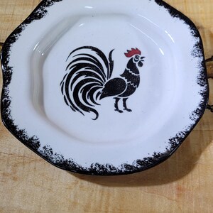 May include: White ceramic plate with a black rooster illustration. The plate has a black, distressed edge. The rooster is black with a red comb. The plate is on a light wood surface.