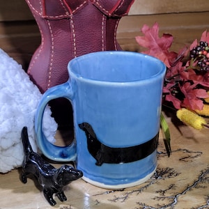 May include: A light blue ceramic mug with a black dachshund design. A small black dachshund figurine sits nearby. A burgundy leather bust and decorative tray with a tree-like pattern are in the background.