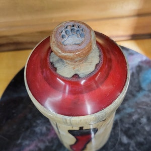 May include: A wooden bottle stopper with a red top and a bear paw print on the knob. The bottle stopper is made of natural wood and has a rustic look.