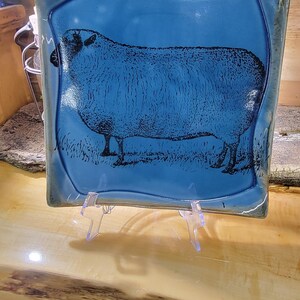May include: Square ceramic plate with a blue glaze and a black sheep illustration. The sheep is standing on grass. The plate is displayed on a clear plastic stand.