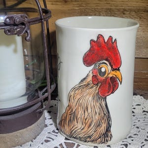 May include: White ceramic mug featuring a detailed illustration of a rooster's head with a red comb and wattle, and a yellow beak. The mug is set on a white lace doily, with a rustic lantern in the background.