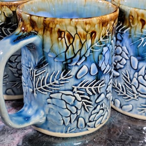May include: Handmade ceramic mugs with a blue and brown glaze. The mugs feature a raised, textured design of leaves and floral patterns. The handles are a matching blue. The mugs are suitable for hot or cold beverages.