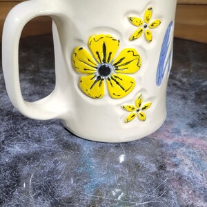 May include: Cream-colored ceramic mug with a large handle, decorated with yellow flowers and blue accents. The flowers have black details. A charming and cheerful design for a coffee or tea cup.