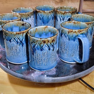 May include: A collection of blue ceramic mugs with a mountain and forest design. The mugs have a textured surface and a brown rim. The mugs are displayed on a round, dark-colored tray.