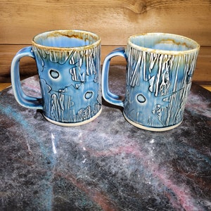 May include: Two blue ceramic mugs with a white and blue patterned design. The mugs have a brown rim and a handle.