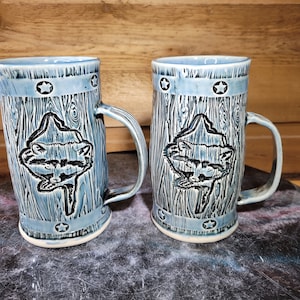 May include: Two blue ceramic mugs with a raccoon design and white stars around the rim. The mugs have a wood grain texture.