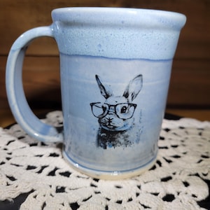 May include: A light blue ceramic mug with a white rabbit wearing glasses design. The mug has a white handle and a textured glaze.