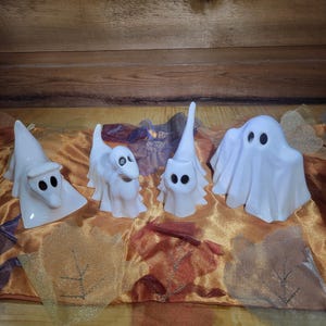 May include: Four white ceramic ghost figurines with black eye details are arranged on a gold and brown patterned surface. The ghosts have different shapes and sizes, perfect for Halloween decor.