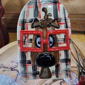 May include: A ceramic decorative cutting board with a plaid pattern in black, red, and white. The board features a cartoon dog face with red glasses. The board has a handle at the top.