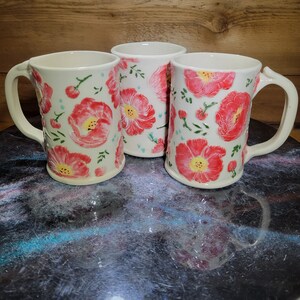 May include: Three white ceramic mugs with a floral pattern. The mugs have a pink and white floral design with green leaves. The mugs are on a black surface.