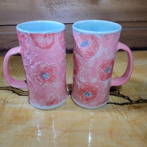 May include: Two pink ceramic mugs with a floral design. The mugs have a light blue rim and a white interior. The floral design features pink flowers with white centers and green leaves.