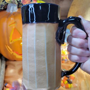 May include: A tall, ceramic mug with a black rim and handle. The mug has a light brown glaze with vertical white stripes. The mug is sitting on a table with candy corn scattered around it.