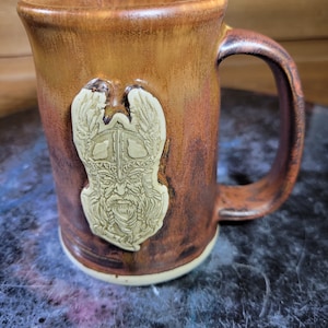 May include: A brown ceramic mug with a white relief design of a Viking warrior with wings. The mug has a handle and a white rim.