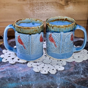 May include: Two ceramic mugs with blue exteriors and handles, featuring a red cardinal bird design. The rims are a darker brown, and the mugs sit on a white lace doily. The background is a wooden surface.
