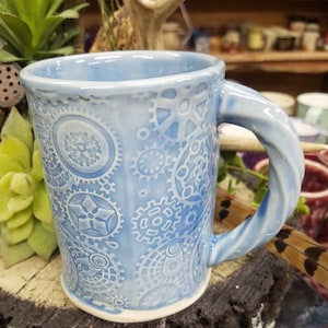 May include: A light blue ceramic mug with a white embossed design of gears and cogs. The mug has a twisted handle.