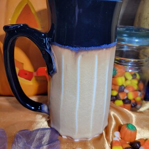 May include: A black and tan ceramic mug with a white stripe design. The mug has a wide, rounded handle and a tall, cylindrical shape.