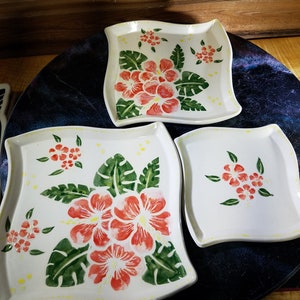 May include: Three white ceramic square plates with scalloped edges, decorated with green leaves and red hibiscus flowers. The plates are on a dark blue background.