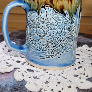 May include: A ceramic mug with a light blue glaze and a brown rim. The mug features a detailed relief design of a bird in a nest with eggs. The handle is light blue. The mug is sitting on a white lace doily.