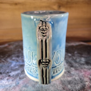 May include: A light blue ceramic mug with a skull design on the handle. The mug has the text "Blue You" on the front and "Treats" on the back.