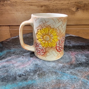 May include: Cream-colored ceramic mug with a sunflower and rose design. The mug features a yellow sunflower in the center and red roses on the sides. The background has a woven basket pattern.