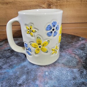 May include: Cream-colored ceramic mug decorated with yellow and blue floral designs. The mug has a curved handle and is set against a wooden background. A decorative item for the kitchen.