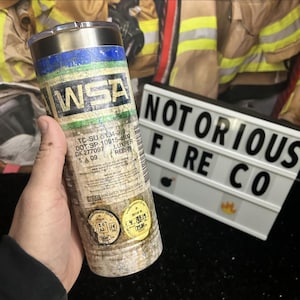 May include: A stainless steel tumbler with a weathered, distressed look. The tumbler features a faded green, blue, and white logo that reads "WSA". The tumbler also has a label with the text "TC-SU 5134-310, DOT-SP-10915-4500, OK 277097, LUXPER, REE: 78, 5A 09".