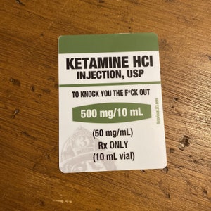 May include: A white and green rectangular sticker with the text "KETAMINE HCI INJECTION, USP" and "TO KNOCK YOU THE F*CK OUT". It also includes "500 mg/10 mL" and "Rx ONLY". The sticker has a logo and the website NotoriousLB3.com.