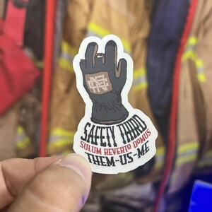 Puede incluir: Una pegatina blanca con una ilustración en negro y marrón de una mano enguantada. La pegatina tiene el texto "Safety Third" en una fuente en blanco y negro. Debajo del texto está la frase "Sulum Revert to Domus" en rojo. El texto "Them-Us-Me" está en negro.