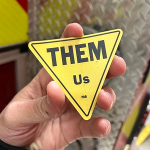 Puede incluir: Un letrero triangular amarillo con las palabras "Them", "Us" y "Me" impresas en negro.