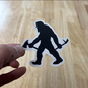 Puede incluir: Pegatina de silueta negra de un Bigfoot con un casco de bombero y sosteniendo un hacha.
