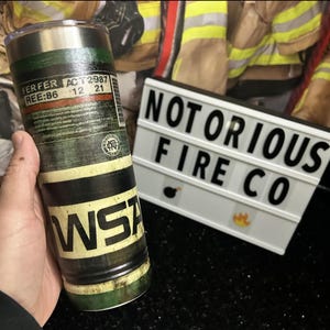 May include: A stainless steel tumbler with a green, black, and white design. The tumbler has the text "FER FER ACT 2387 REE: 86 12 21" and "WSA" printed on it. The tumbler is in front of a white sign with black text that reads "NOTORIOUS FIRE CO".