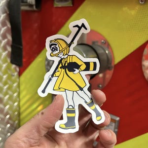Puede incluir: Una pegatina de bombero amarilla y negra con una niña que sostiene una manguera de incendios y lleva un casco con la palabra "消防" en él.
