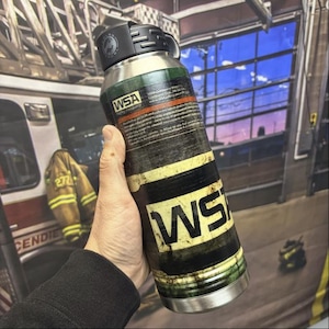May include: A stainless steel water bottle with a black screw-top lid. The bottle features a wrap-around graphic design resembling a weathered green and yellow industrial gas cylinder with black text that reads WSA and Safety Company.